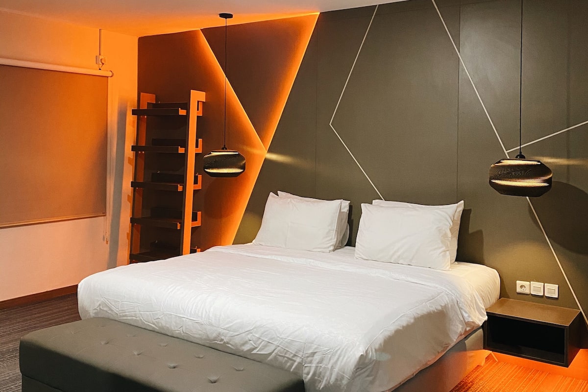 A modern bedroom with a large bed, a bedside table, a bench, and a wall-mounted shelf.