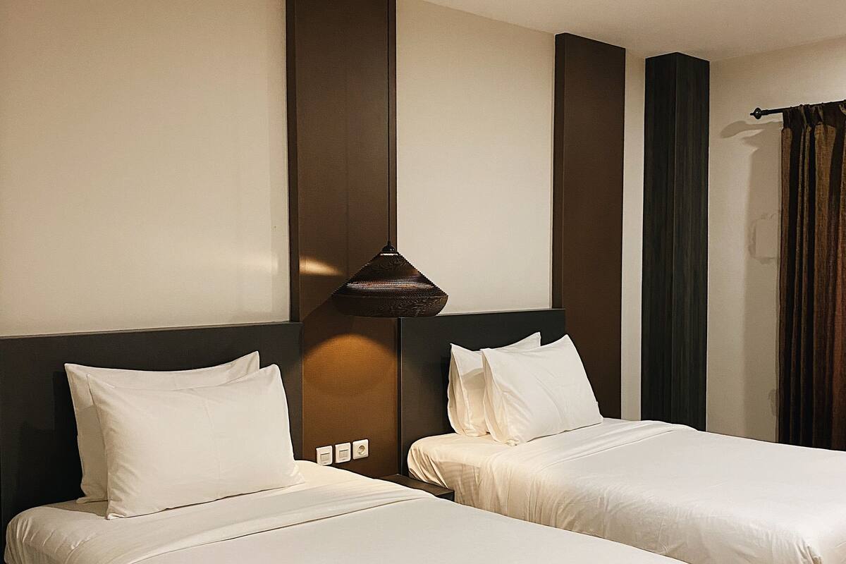 A hotel room with two beds, a dark headboard, a hanging lamp, and a window with curtains.