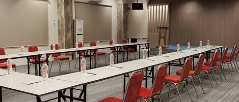 A conference room with long tables, red chairs, water bottles, and a projector screen.