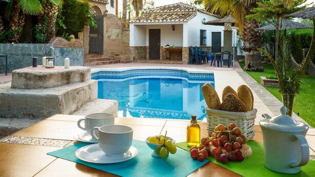 A poolside breakfast setup with a table, two cups, a bowl of grapes, a bottle of oil, and a jug of milk.