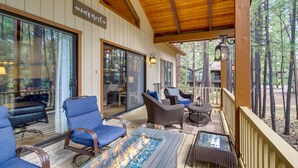 A covered outdoor seating area with blue chairs, a glass-top table with a fire feature, and a view of the surrounding forest.