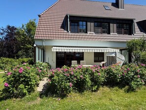 Une maison à deux étages avec un porche couvert, une cheminée et un jardin agrémenté de fleurs roses.