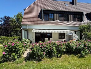 A two-story house with a covered porch, a chimney, and a garden with pink flowers.