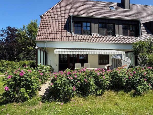 A two-story house with a covered porch, a chimney, and a garden with pink flowers.