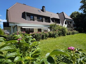A two-story house with a sloped roof, a visible chimney, and a garden with green plants and pink flowers. - Seewolke Vacation Apartment No. 3 - Seewolke No. 1, No. 2, No. 3, Katingsiel near (Tönning)
