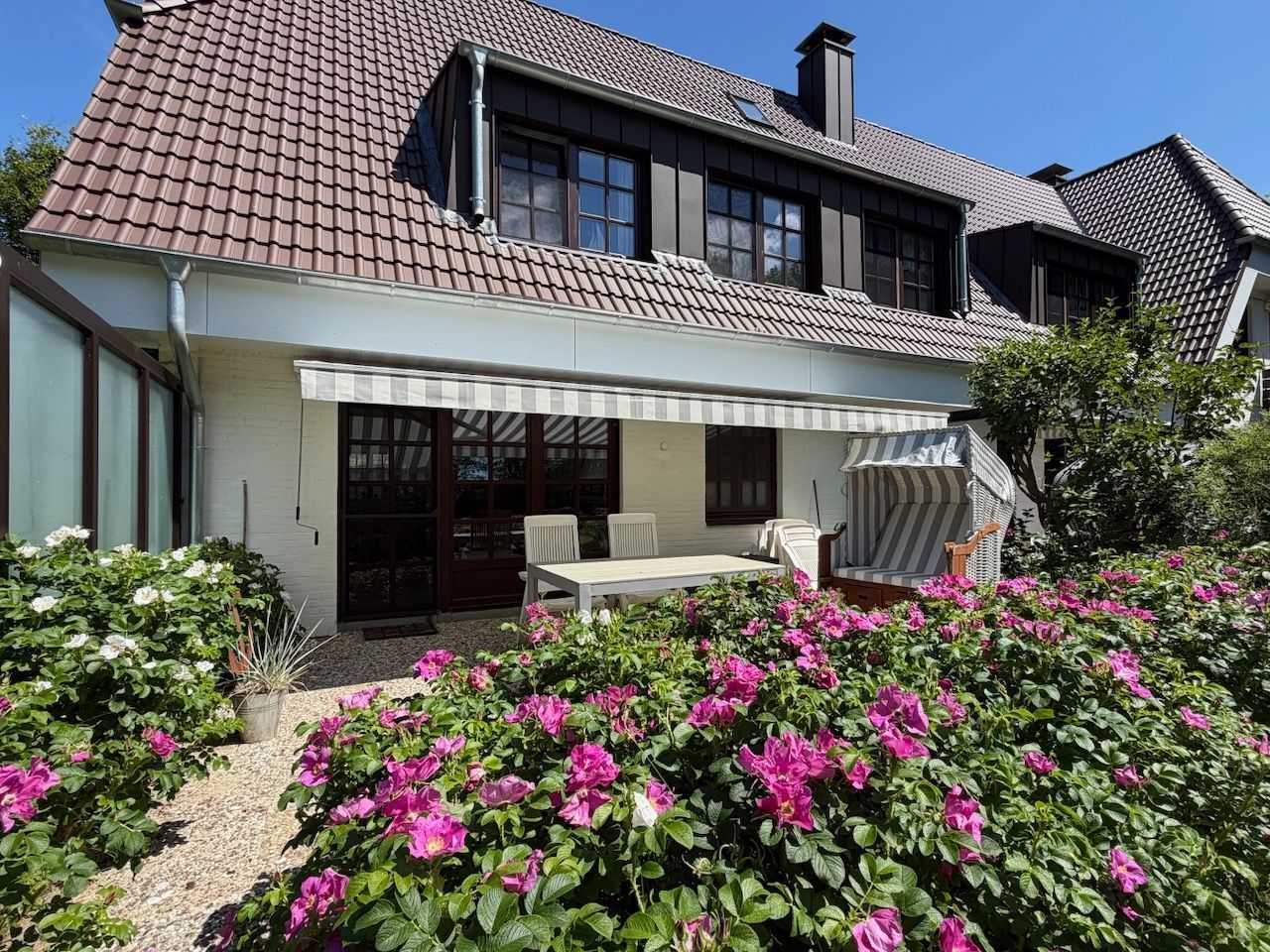 A two-story house with a covered patio, a dining area, and a garden with pink flowers.