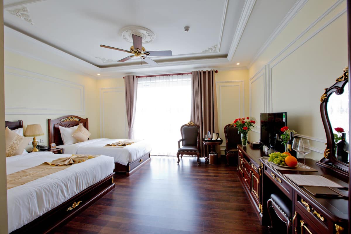 A hotel room with two beds, a ceiling fan, a desk with a TV, a vase with flowers, and a window with curtains.