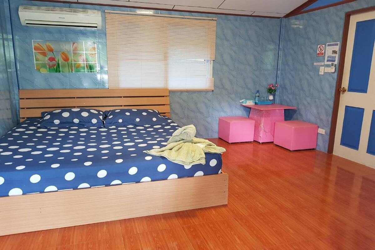 A bedroom with a bed, a window with blinds, an air conditioning unit, a small pink table, and a door.