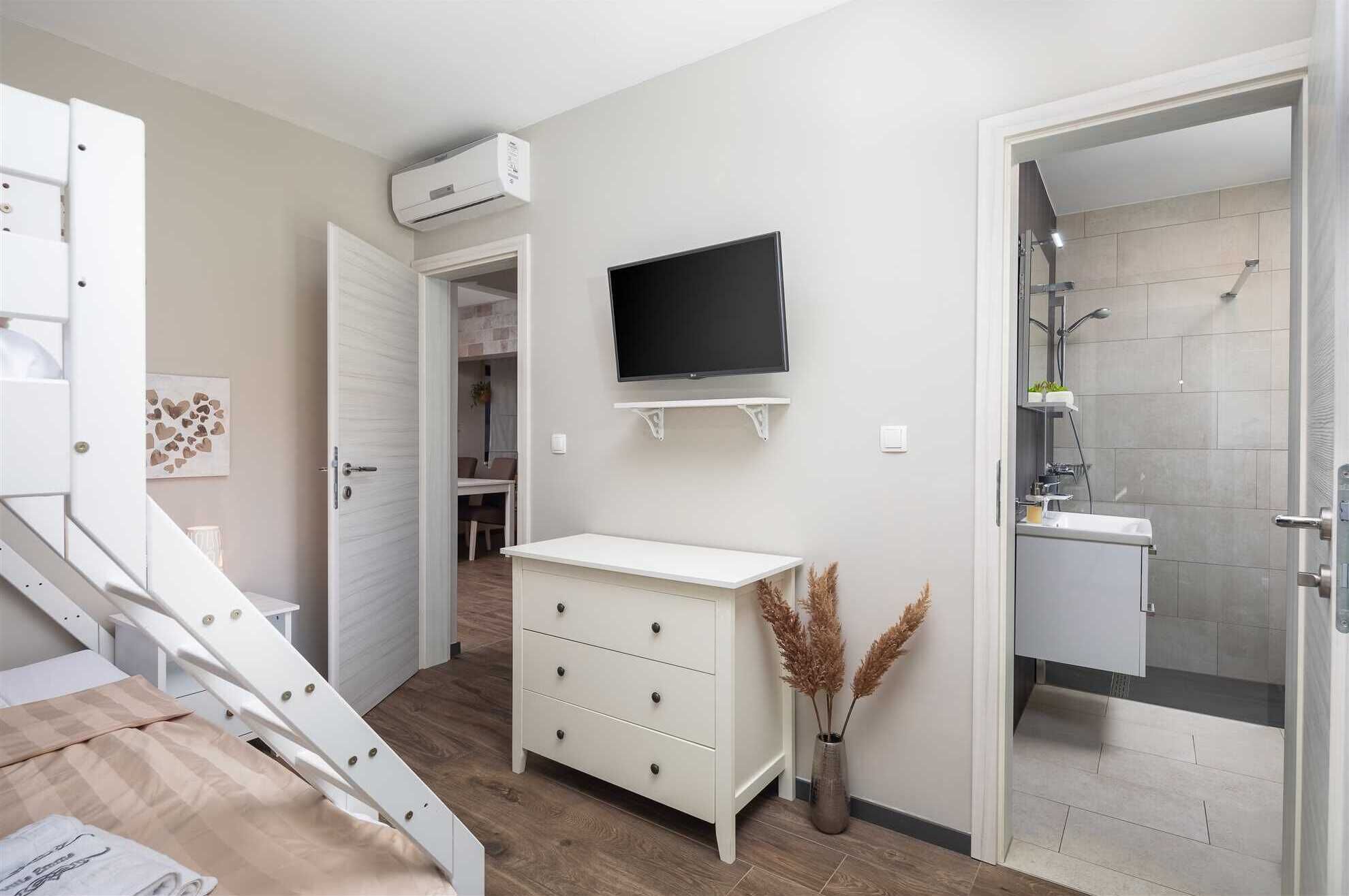 A modern bathroom with a shower, toilet, and sink. There is a white vanity with drawers and a mounted TV above it.