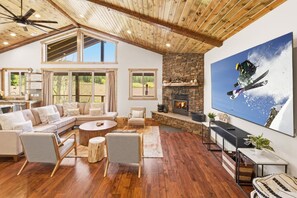 A spacious living room with a stone fireplace, a large TV, and a high ceiling with exposed beams.