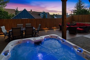 Un jacuzzi en una terraza de madera con mobiliario exterior y vistas a casas al fondo.