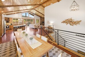 A spacious living area with a wooden dining table, a staircase, and a view of the kitchen. - Located in Moonridge Near Skiing, Golfing, Hiking, & Zoo! Views! Central A/C! HOT TUB! Pool Table, Shuffleboard & Foosball! (Big Bear Lake)