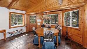 Interior de una cabaña de madera con una mesa redonda para comer, sillas, una araña y ventanas grandes.