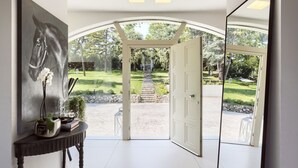 Ein Innenraum mit einem großen Gemälde eines Pferdes, einem Tisch mit einer Pflanze und Blick auf einen Garten durch eine offene Tür.