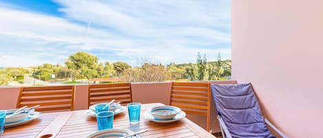 Une table en bois, dressée avec des assiettes, des bols et des verres, une chaise bleue rembourrée, et une vue sur des arbres et des collines.