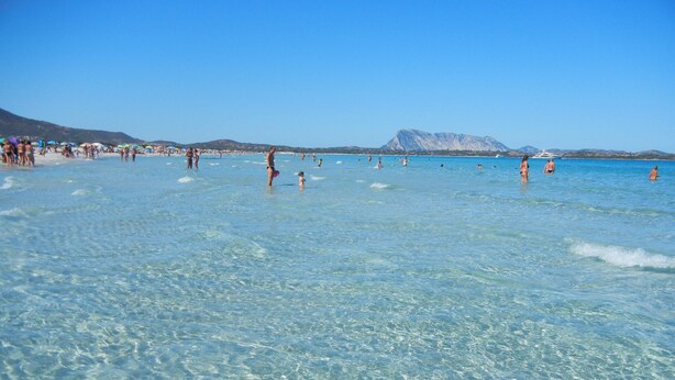 Une plage avec une eau cristalline, des gens qui se baignent et une montagne en arrière-plan.