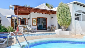 A pool with a tiled edge, a covered patio with chairs, and a building with a door and windows. - Villa Alex with WiFi and Pool (Puerto del Carmen)