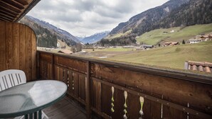 Une terrasse en bois avec une table et des chaises en verre, donnant sur une vallée parsemée de maisons et bordée de montagnes.