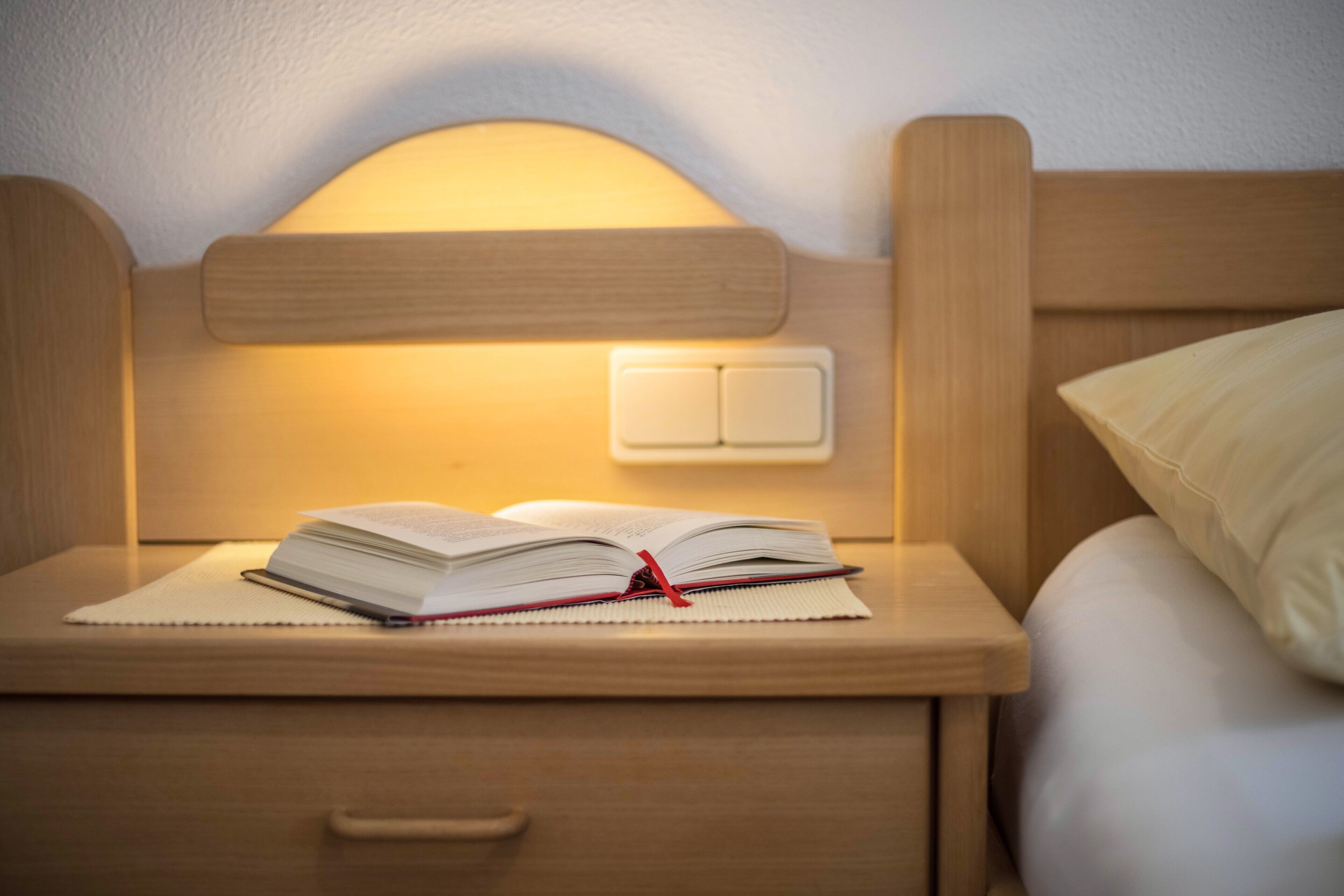 Una cama bien hecha con una almohada, una mesita de noche con un libro abierto y una lámpara de pared.