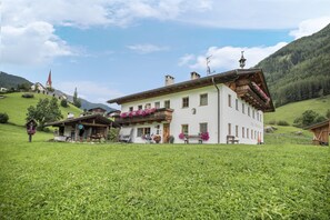 A large white building with a balcony, surrounded by lush green fields and mountains.