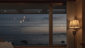 A room with a large window overlooking the sea, a lamp on the right, and a bed with a white pillow. - HOTEL Churabeach (Motobu)