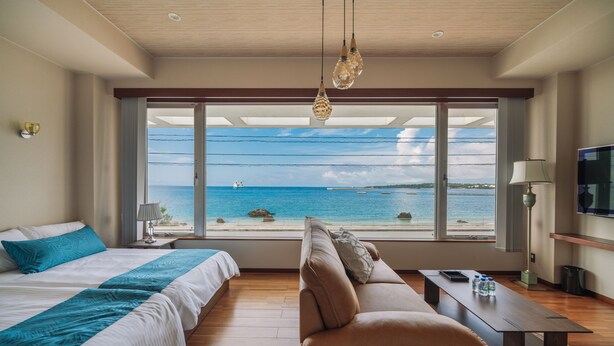 A bedroom with a large window offering a view of the sea, a bed with blue bedding, a brown sofa, and a small table with bottles. - HOTEL Churabeach (Motobu)