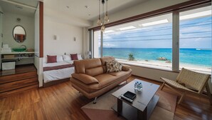 A modern hotel room with a large window offering a view of the ocean, a brown leather sofa, a wooden coffee table, and a small desk with a mirror and shelves. - HOTEL Churabeach (Motobu)