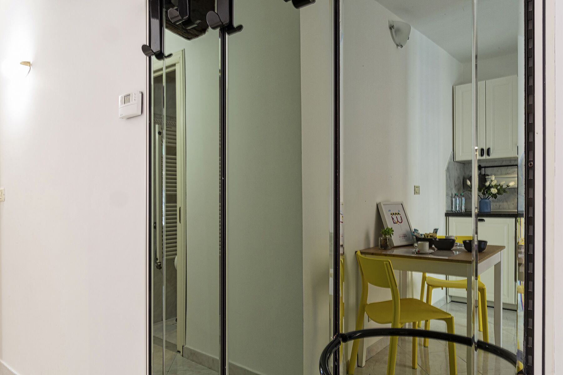 A modern interior with a glass door leading to a bathroom, a dining area with yellow chairs, and a kitchen with white cabinets.