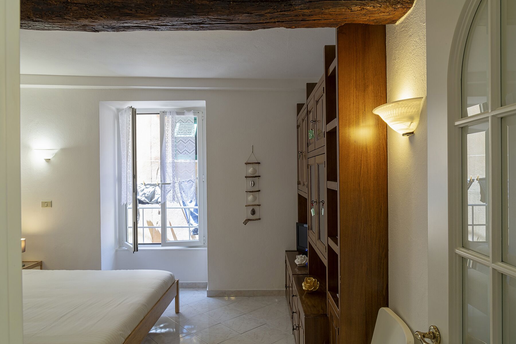 Quarto com cama, janela, guarda-roupa de madeira e prateleira fixada na parede com objetos decorativos.
