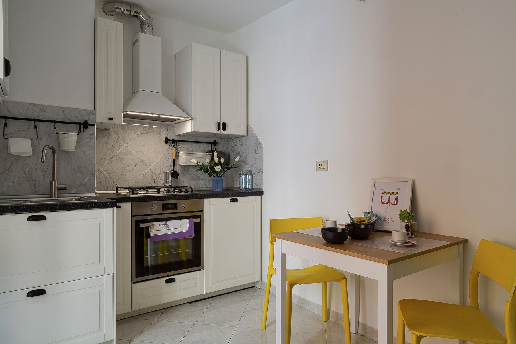 A compact kitchen with white cabinets, a gas stove, a sink, and a small dining table with yellow chairs.