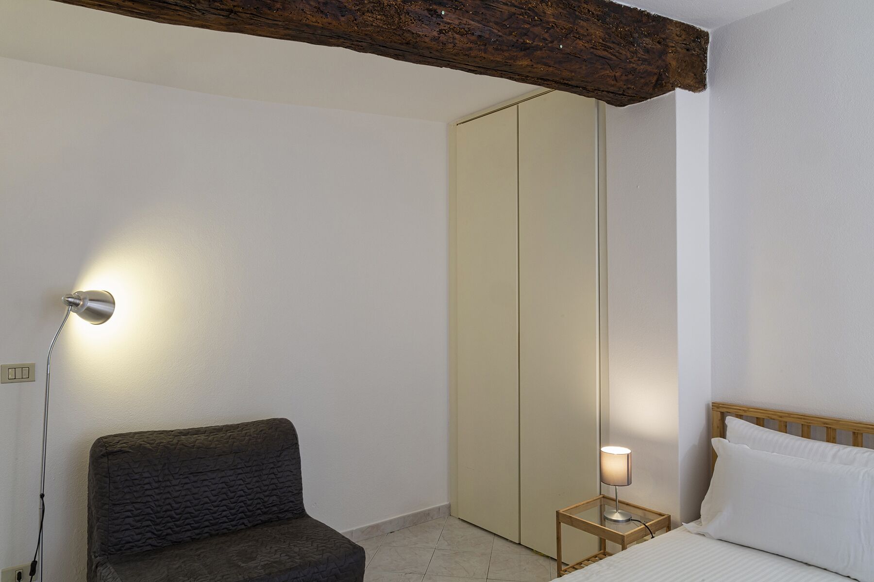 A bedroom with a bed, a bedside table with a lamp, a chair, a wardrobe, and a wooden ceiling beam.