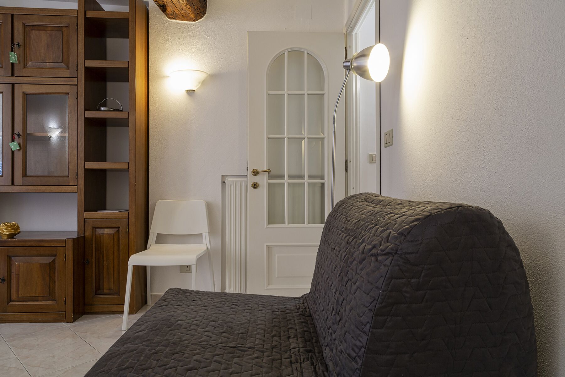 A cozy living room with a dark quilted sofa, wooden cabinets, a white chair, and a white door with a frosted glass window.