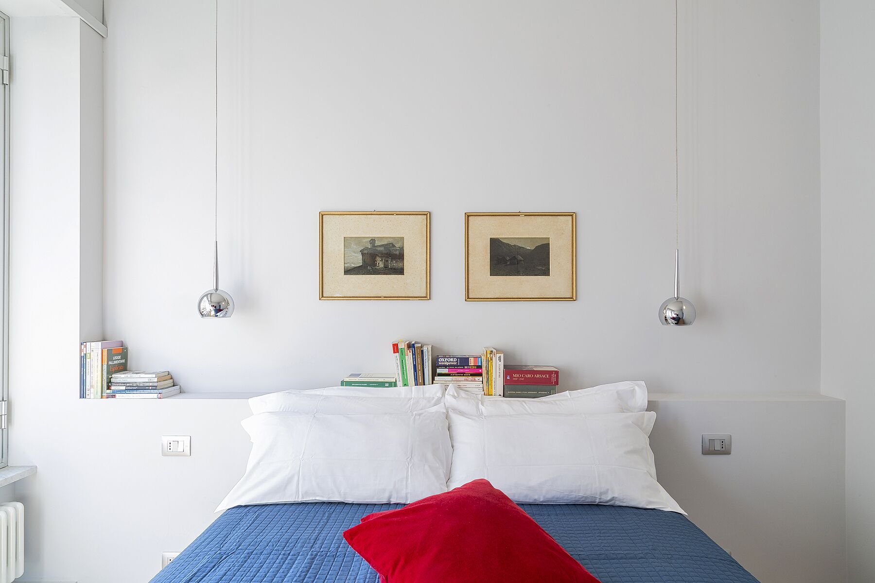 A bedroom with a bed, pillows, a red throw, and books on a shelf.