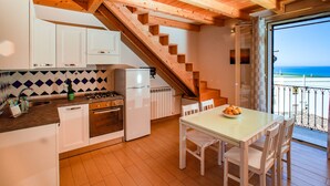 Eine Küche mit weißen Schränken, einem Herd und einem Essbereich mit Tisch und Stühlen. Eine Holztreppe führt zu einem Balkon mit Blick aufs Meer.