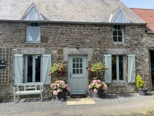 Un bâtiment en pierre avec une porte grise, deux fenêtres avec des volets et un banc à l’extérieur.