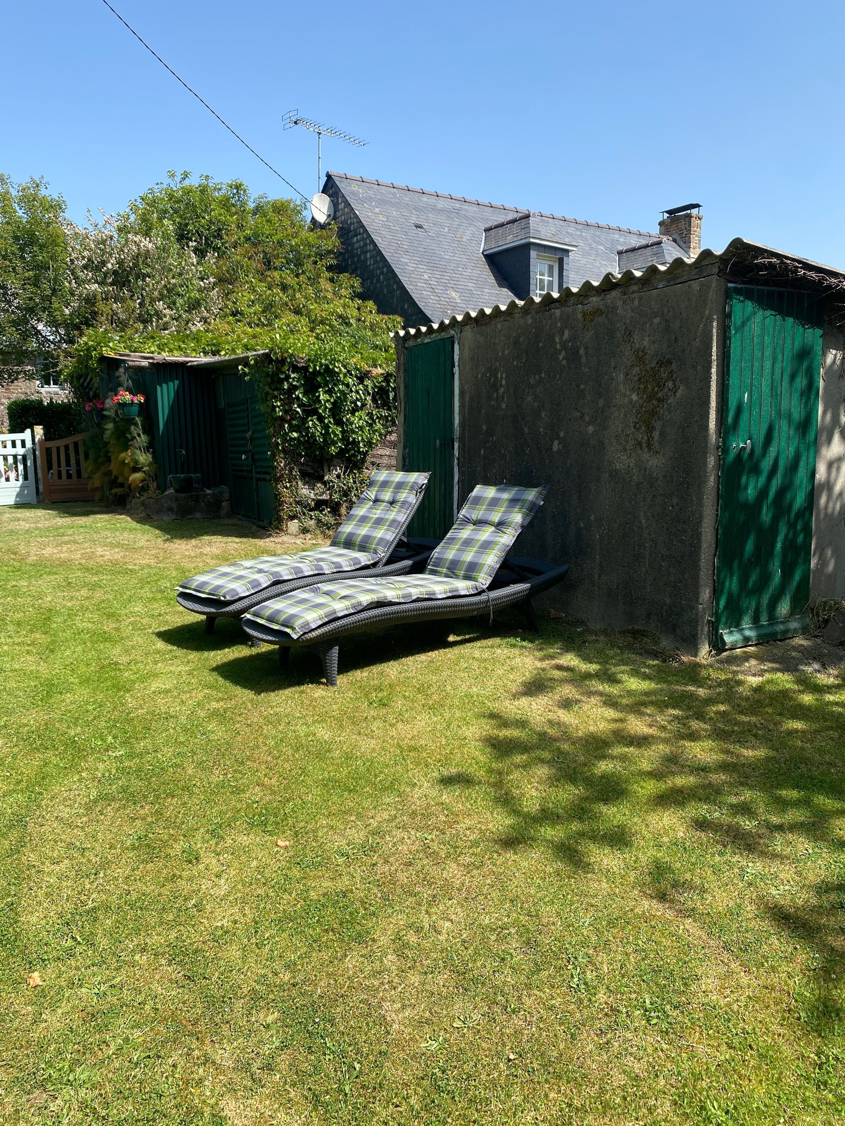 A small, rustic house with a sloped roof and a green door. In front of the house, there is a grassy lawn with two lounge chairs and a small shed.
