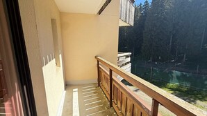A balcony with a wooden railing, a view of a forested area, and a building with a beige wall and a window.