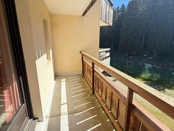 A balcony with a wooden railing, a view of a forested area, and a building with a beige wall and a window.