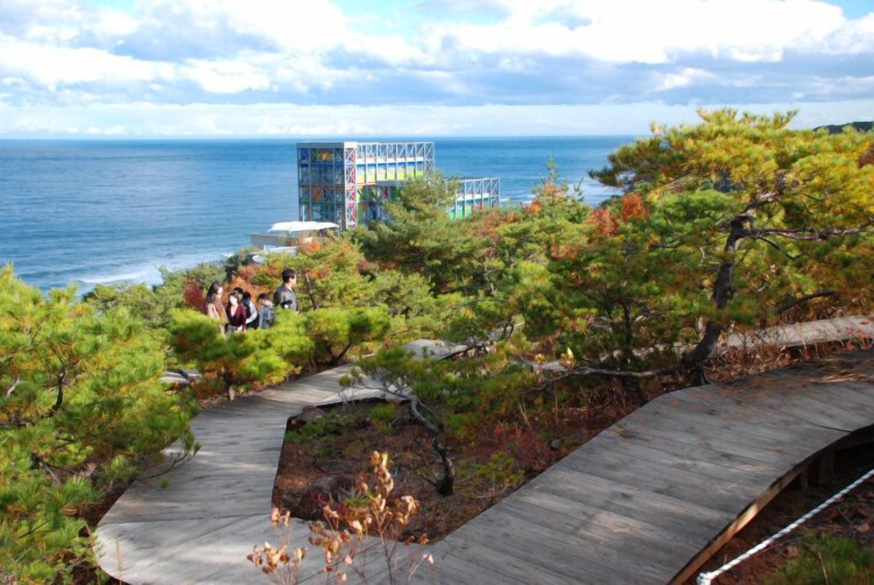 A coastal view with a wooden walkway, a building with colorful exterior, and a group of people.