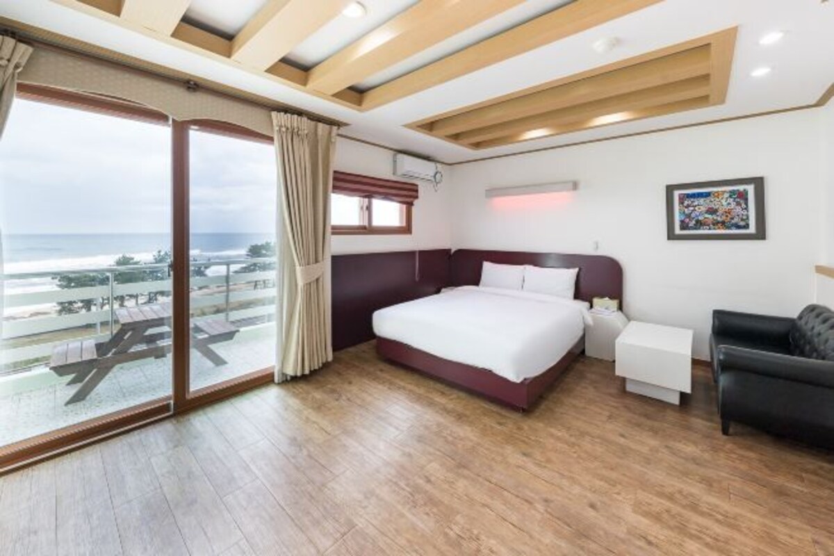 A bedroom with a bed, a nightstand, a black leather chair, a window with curtains, and a view of the beach.