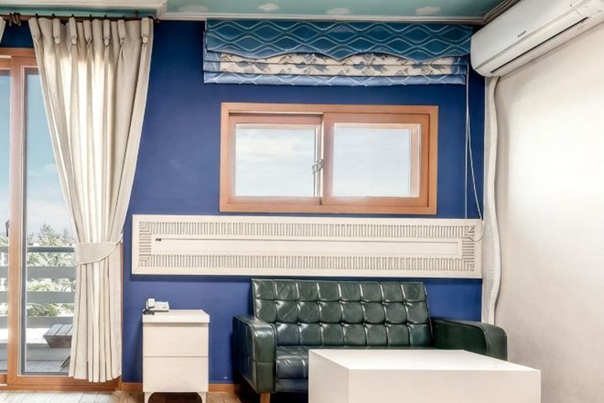 A room with a green leather sofa, a white bedside table, a window with a wooden frame, and a radiator.
