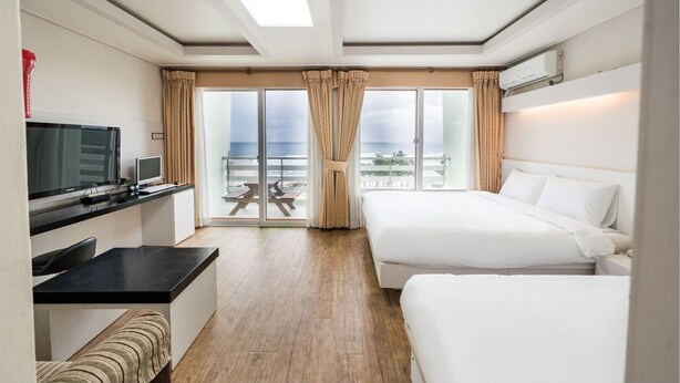 Une chambre d’hôtel avec un lit, un bureau doté d’une télévision et une vue sur l’océan grâce à de grandes fenêtres.