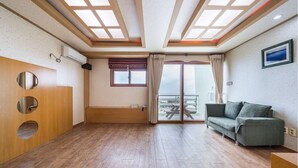 Une pièce spacieuse avec un plancher en bois, un canapé vert, un meuble en bois doté d’ouvertures circulaires, une fenêtre avec des rideaux et une vue sur l’extérieur à travers une porte vitrée.