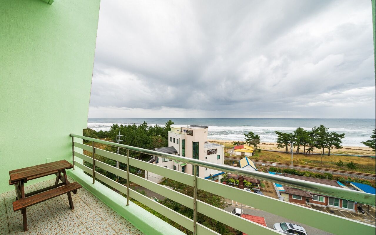 Ein Balkon mit einer hölzernen Bank, der Blick auf einen Strand mit Gebäuden und einem bewölkten Himmel geht.