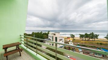 A balcony with a wooden bench, overlooking a beach with buildings and a cloudy sky.
