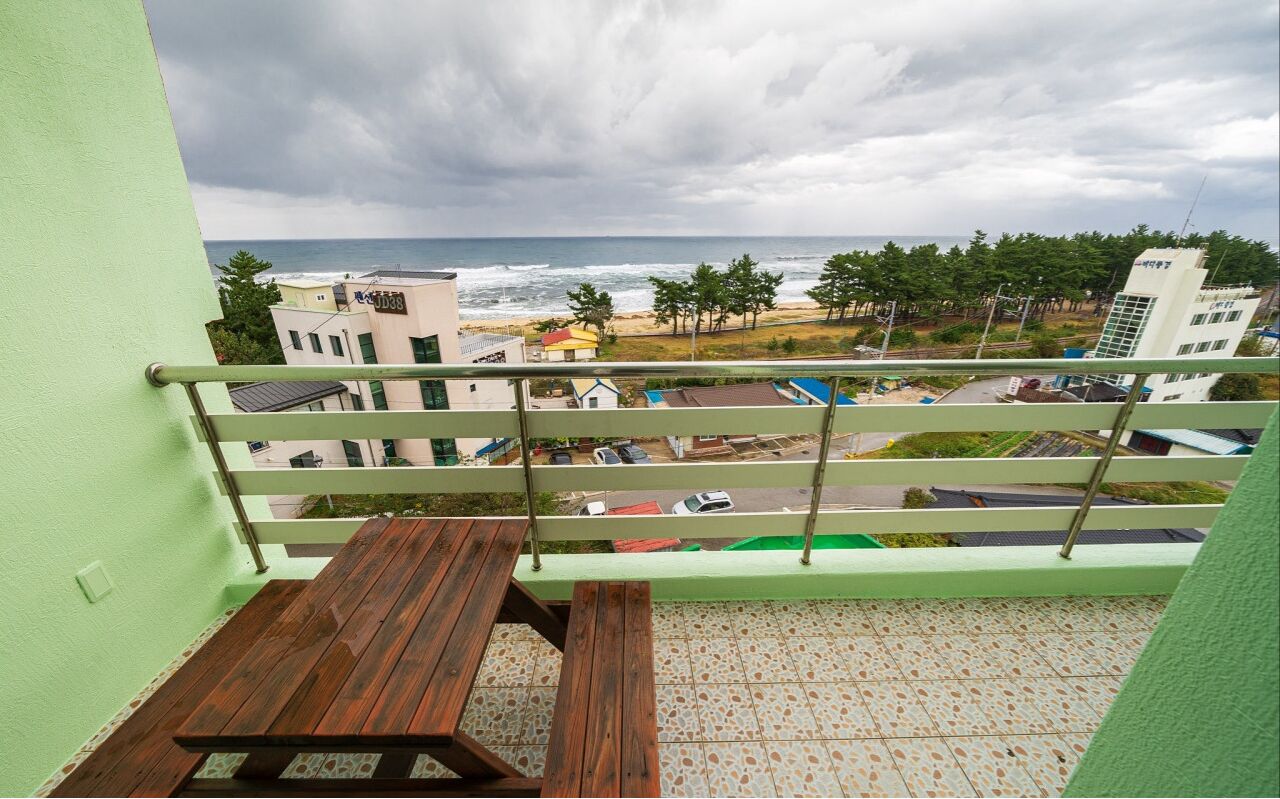 Un balcon doté d’une table en bois et d’une rambarde en métal, offrant une vue sur une plage et le paysage urbain.