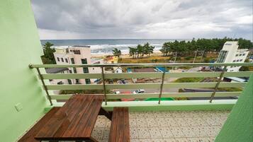 Un balcon doté d’une table en bois et d’une rambarde en métal, offrant une vue sur une plage et le paysage urbain.