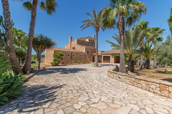 A stone-paved courtyard with a brick house, palm trees, and a stone wall.