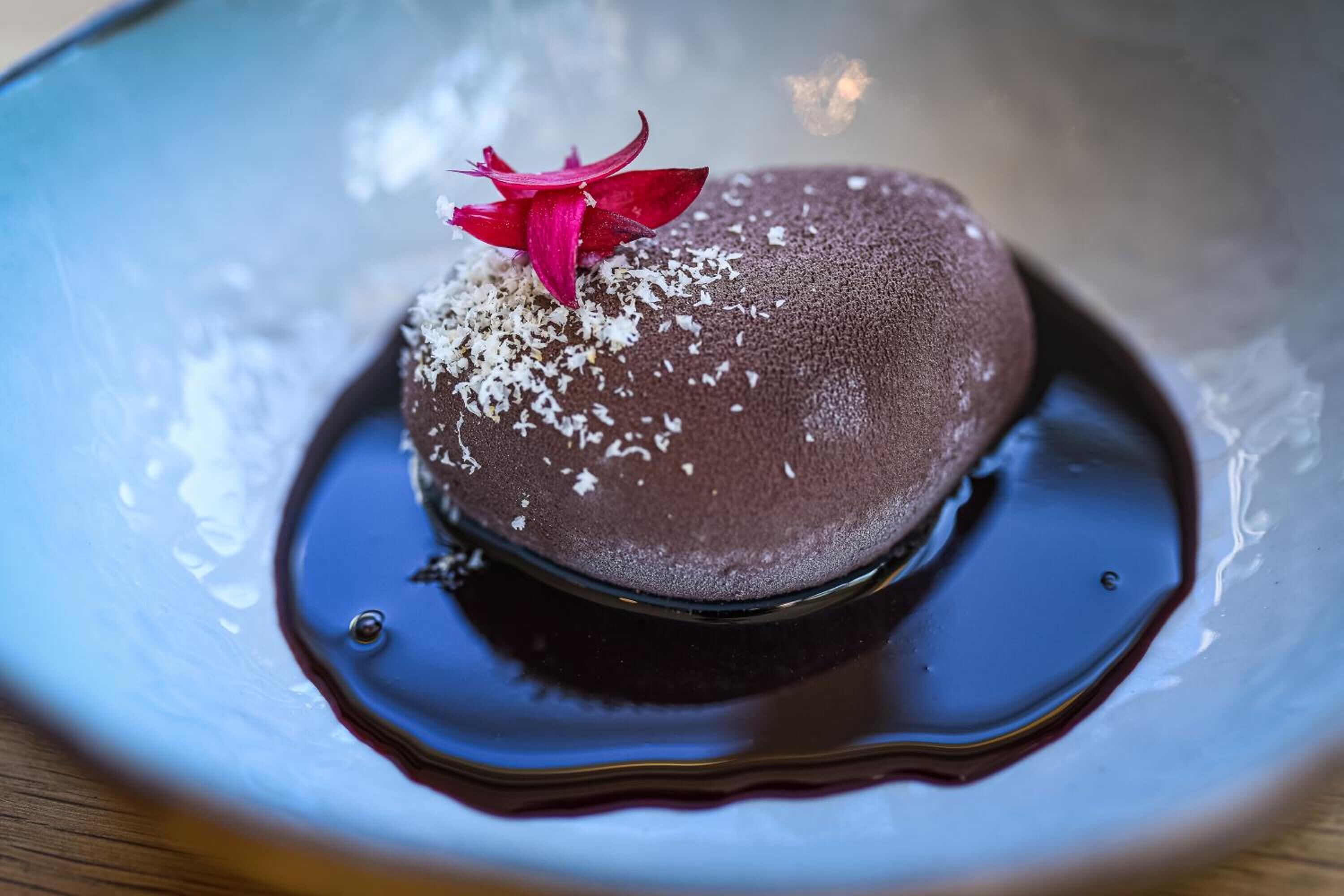 A dessert on a plate with a dark chocolate base, white chocolate shavings, and a pink flower garnish.