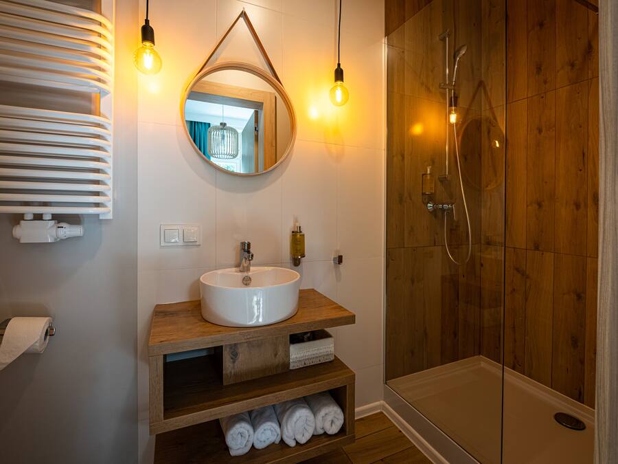 A modern bathroom with a wooden vanity, a round mirror, a white sink, and a glass-enclosed shower.